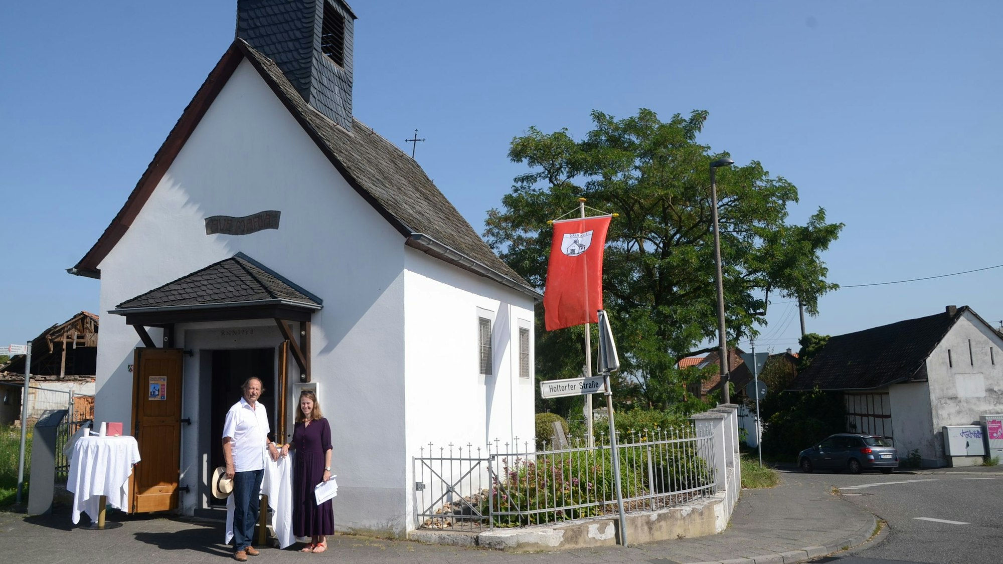 Eine weiße Kapelle mit einem Vordach und einem kleinen Glockenturm. Rechts weht eine rote Flagge mit dem Wappen von Vinxel, dahinter steht ein großer Baum.