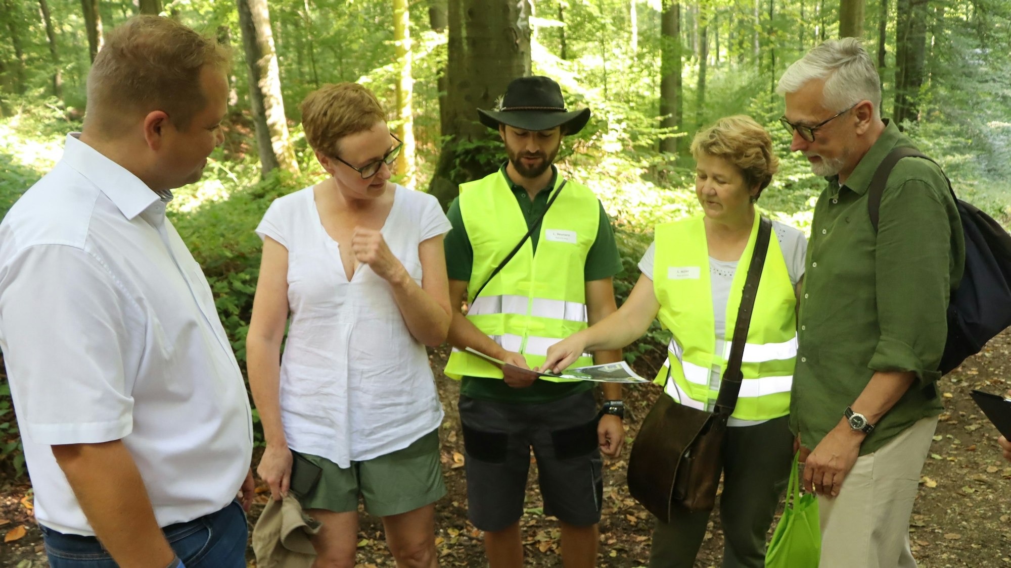 Eine Gruppe aus drei Männern und zwei Frauen, zwei Personen in gelben Warnwesten, schauen im Wald auf eine Karte.