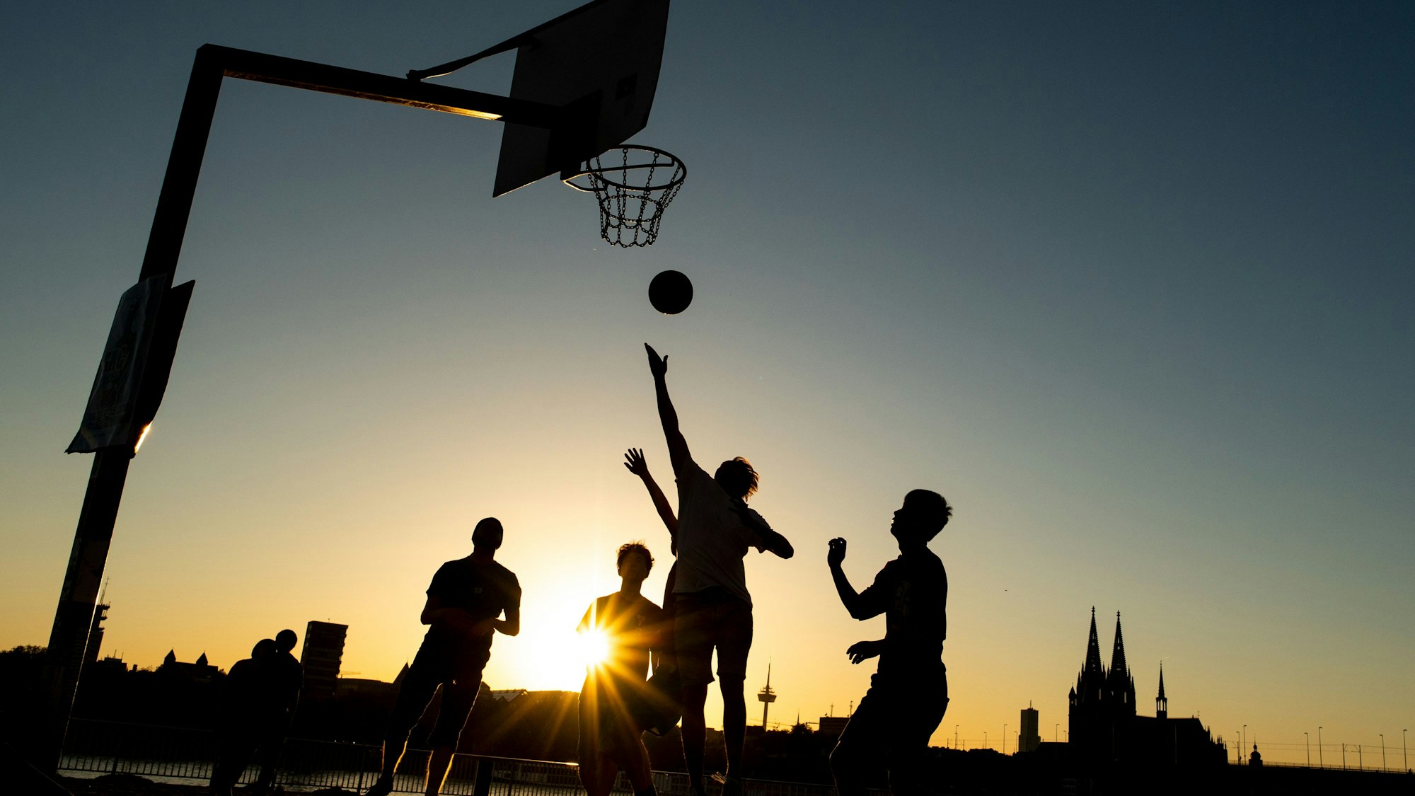 Männer spielen im Licht der untergehenden Abendsonne Basketball am Rheinufer.