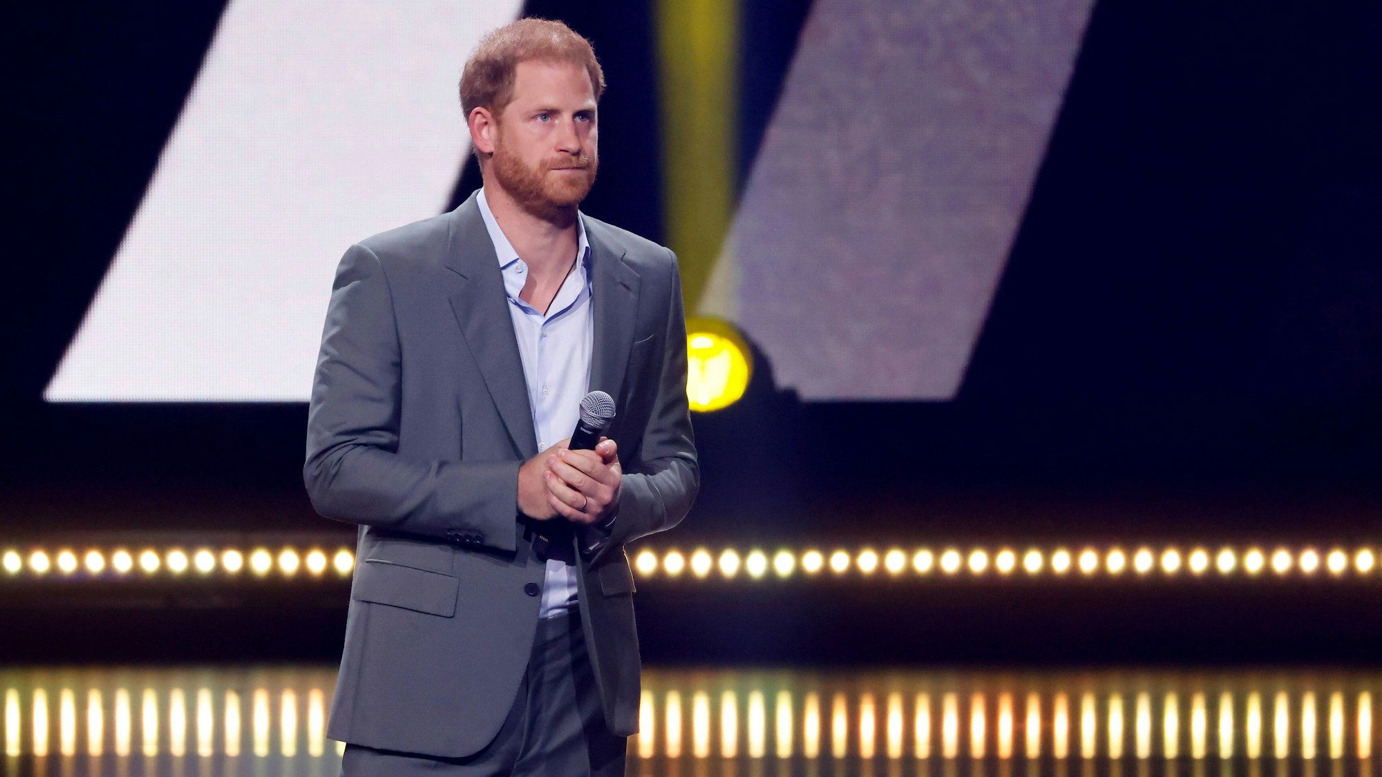 Prinz Harry steht auf der Bühne und presst die Lippen zusammen. Das Mikrofon hält er in beiden Händen.