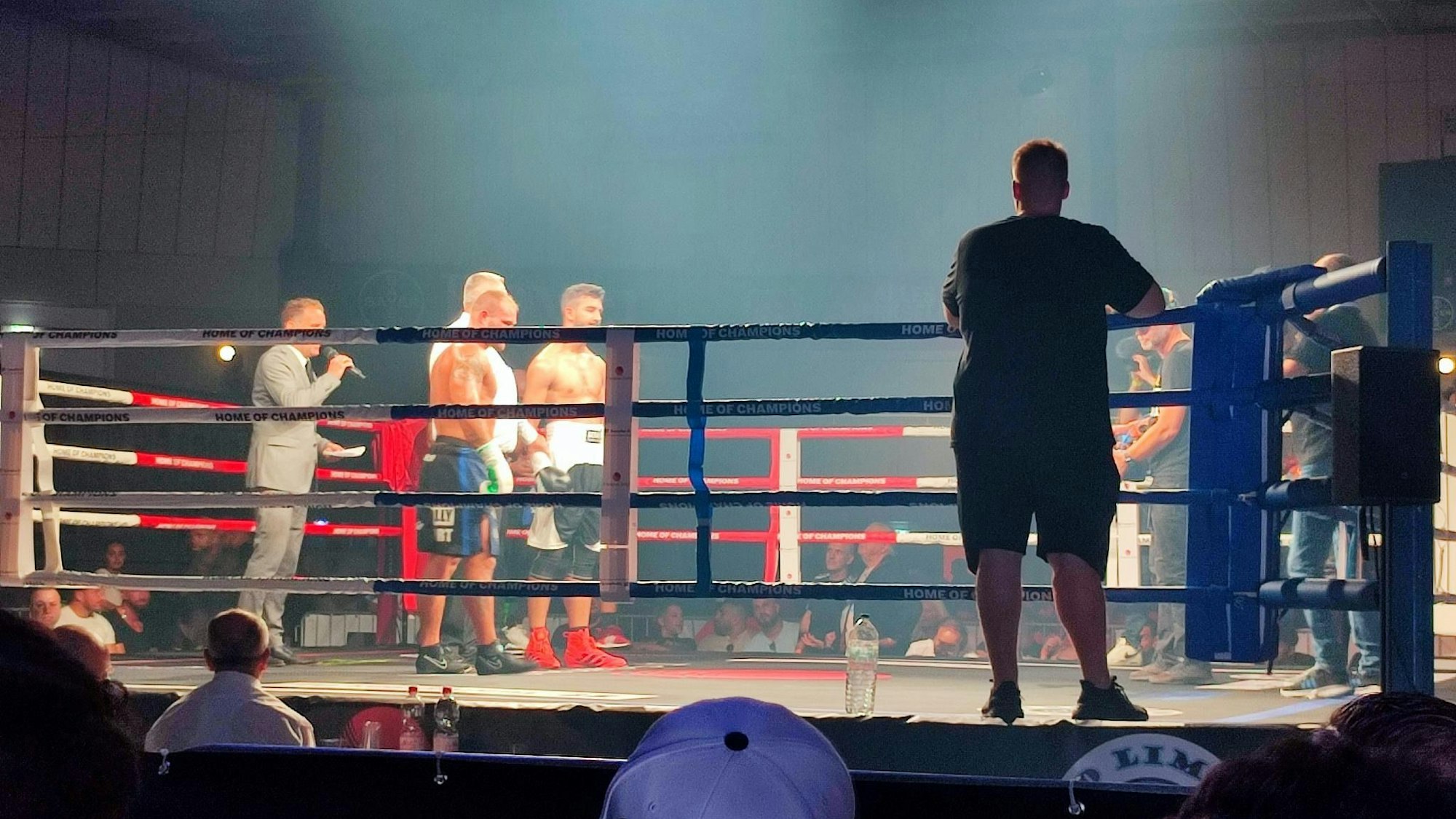Der Ringsprecher verliest das Ergebnis der Jury nach einem Boxkampf. Die Boxer warten auf das Ergebnis, der Ringrichter hält ihre Arme. Im Vordergrund ist Publikum zu sehen.