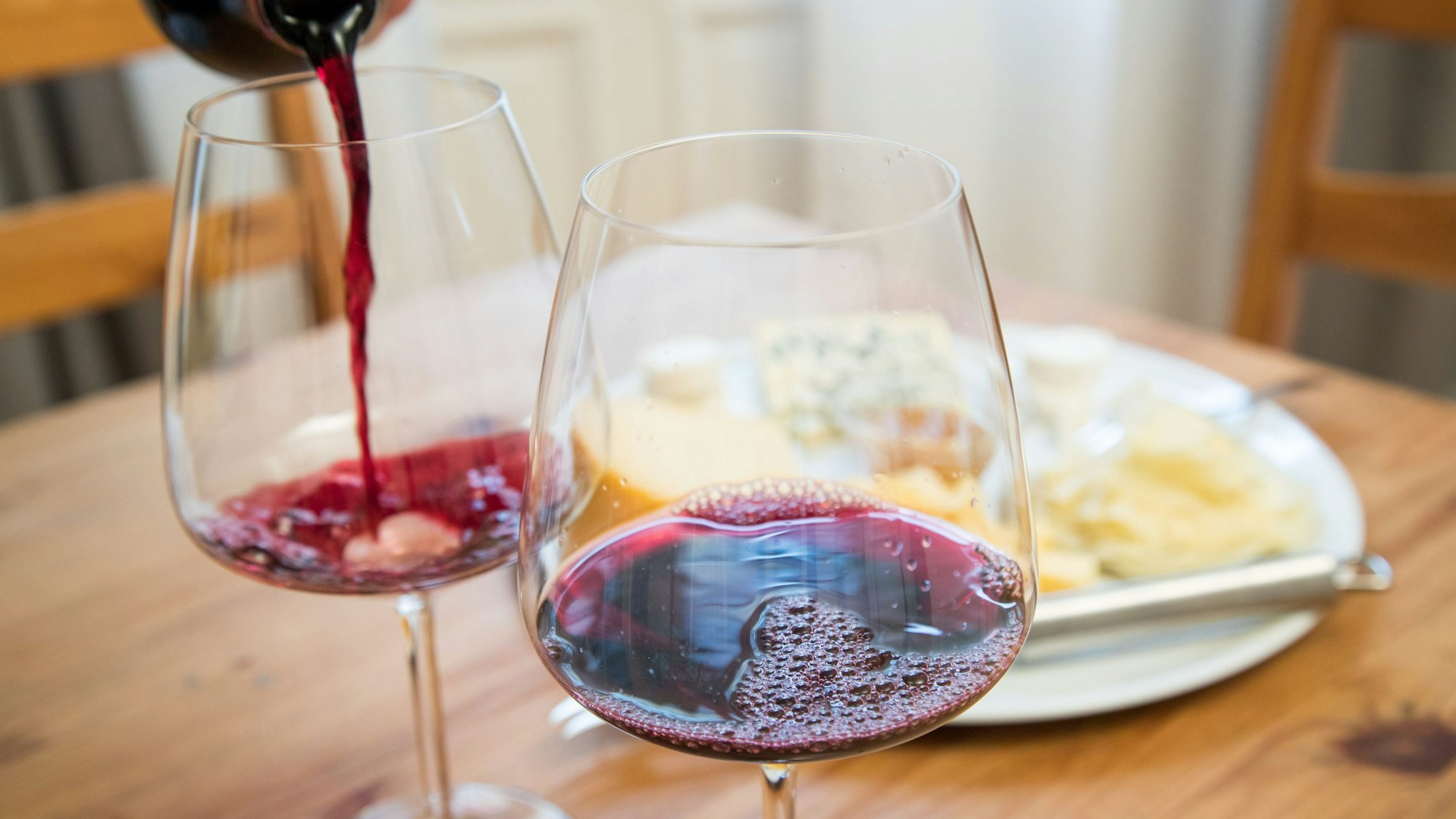 Im Vordergrund sehen zwei Rotweingläser, in eines wird gerade eingeschenkt. Im Hintergrund sieht man unscharf einen Teller mit Essen.