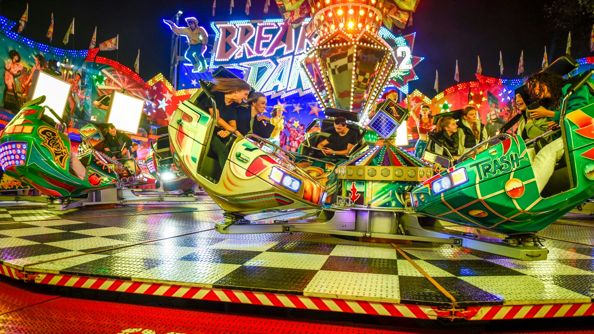 Auf dem Breakdance-Fahrgeschäft wirbeln Menschen in ihren Sitzkabinen hin und her. Das Fahrgeschäft leuchtet in bunten Farben und Lichtern.
