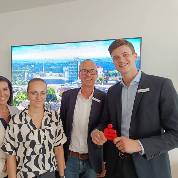 Zwei Frauen und zwei Herren lächeln vor einem großen Fernseh-Bildschirm in die Kamera.