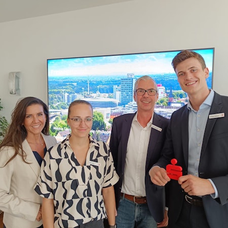 Zwei Frauen und zwei Herren lächeln vor einem großen Fernseh-Bildschirm in die Kamera.