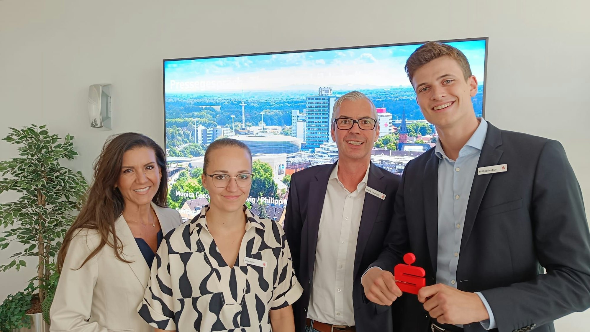 Zwei Frauen und zwei Herren lächeln vor einem großen Fernseh-Bildschirm in die Kamera.