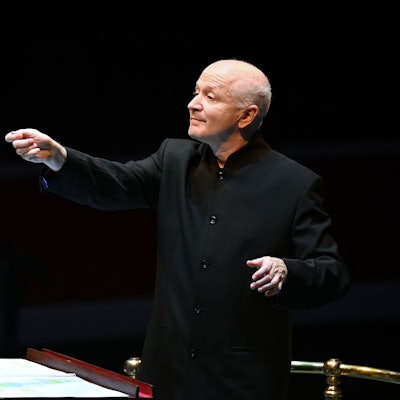 George Benjamin trägt ein schwarzes Sakko und dirigiert, der Hintergrund ist dunkel.