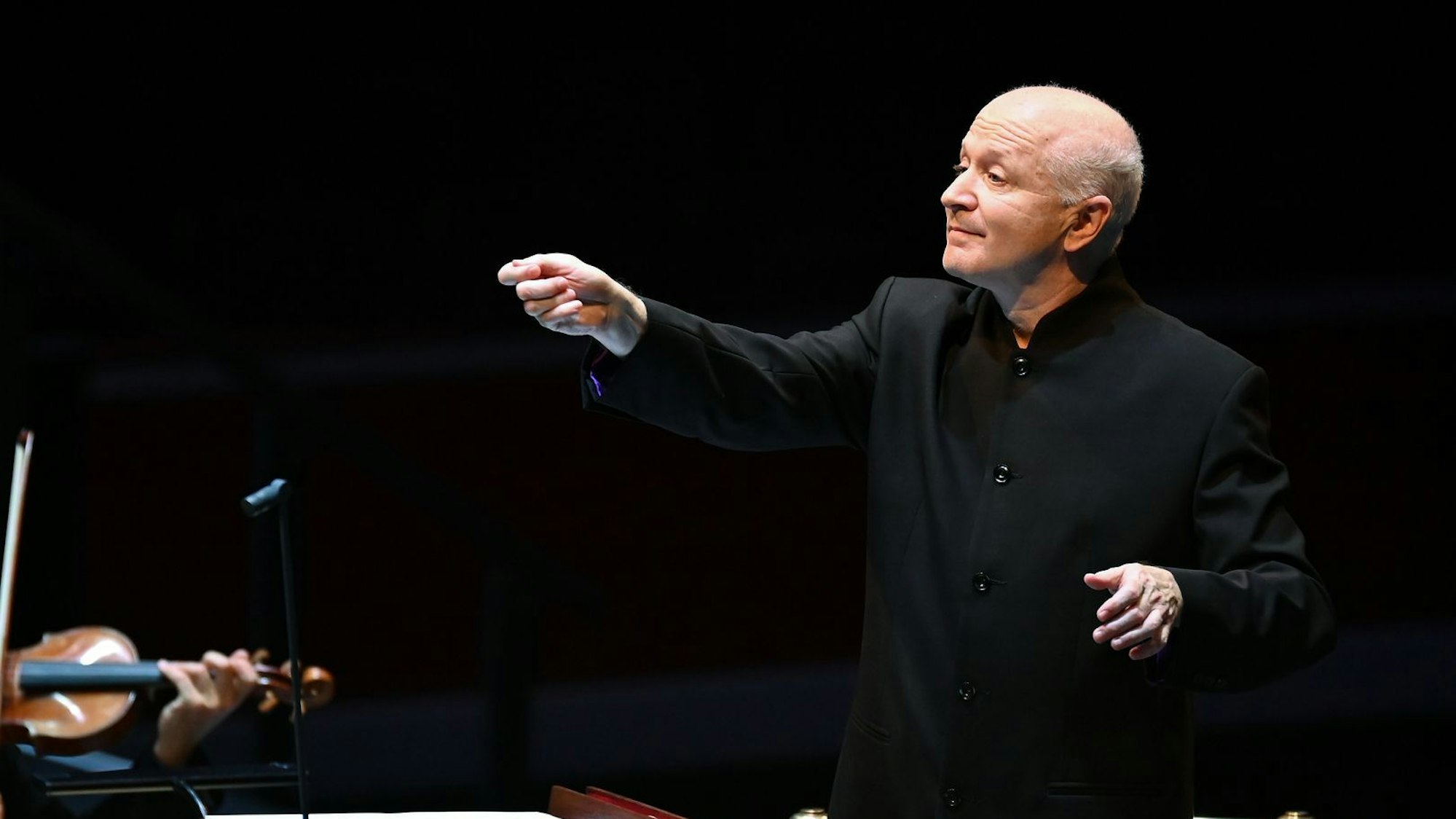 George Benjamin trägt ein schwarzes Sakko und dirigiert, der Hintergrund ist dunkel.