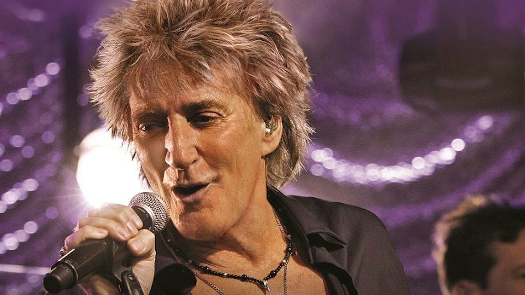 Zu sehe ist der Sänger Rod Stewart, welcher ein Mikrofon in der Hand hält.