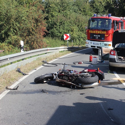 Ein schwer beschädigtes Motorrad liegt auf der Straße, dahinter stehen ein Auto sowie Einsatzfahrzeuge der Feuerwehr Kürten.