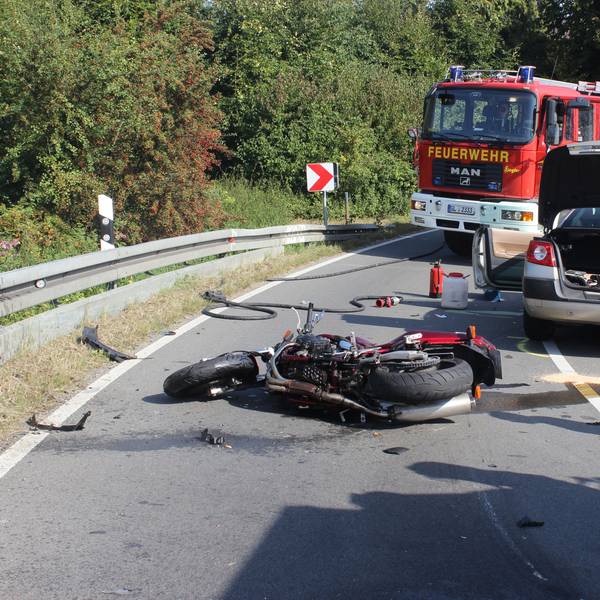 Ein Motorrad liegt auf der Fahrbahn, dahinter der Unfall-Pkw und zwei Einsatzfahrzeuge der Feuerwehr.