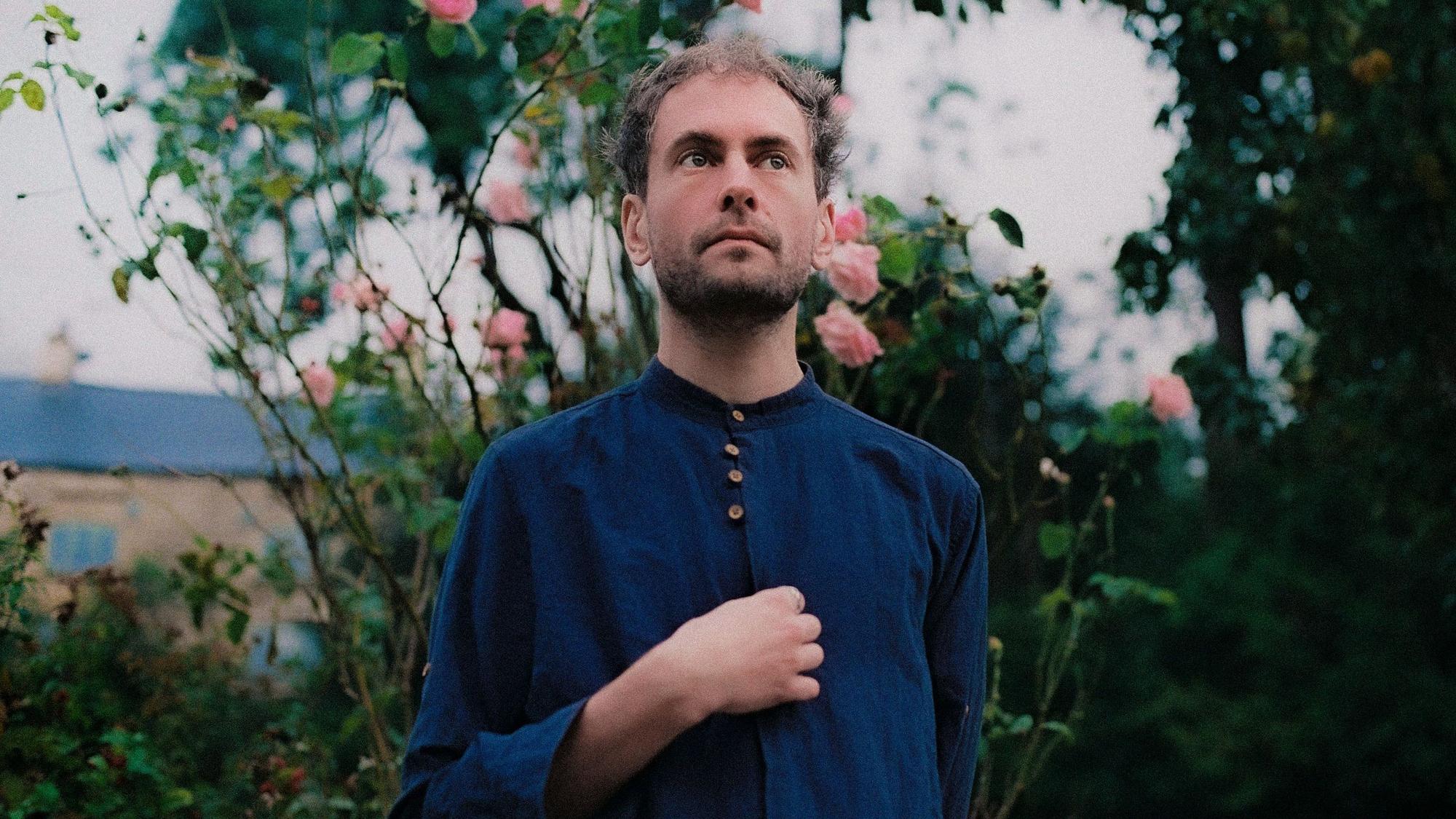 Alabaster DePlume trägt ein blaues zugeknöpftes Hemd. Im Hintergrund ist ein Rosenbusch.