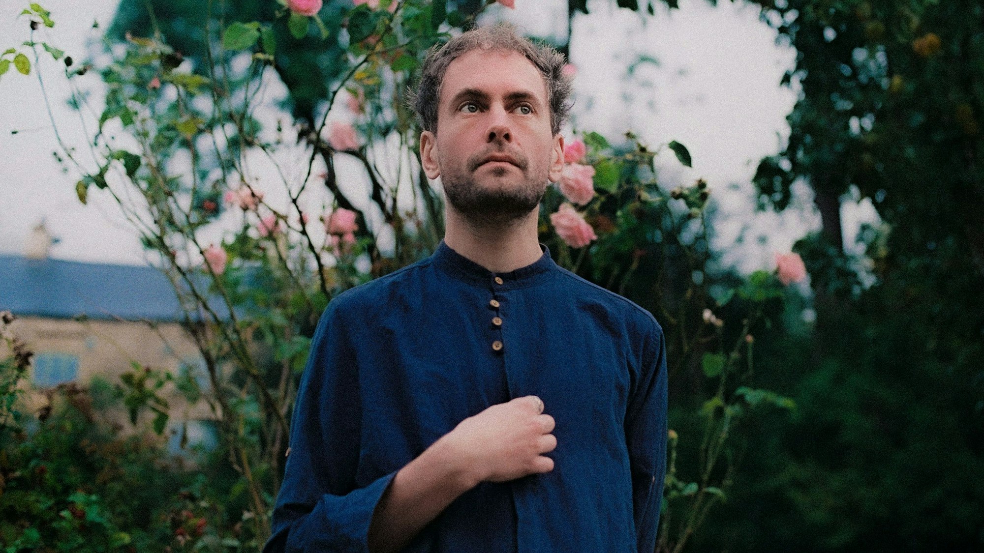 Alabaster DePlume trägt ein blaues zugeknöpftes Hemd. Im Hintergrund ist ein Rosenbusch.