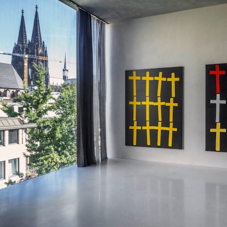 Zwei Bilder mit Kreuzen hängen neben einem Fenster, durch das der Kölner Dom zu sehen ist.