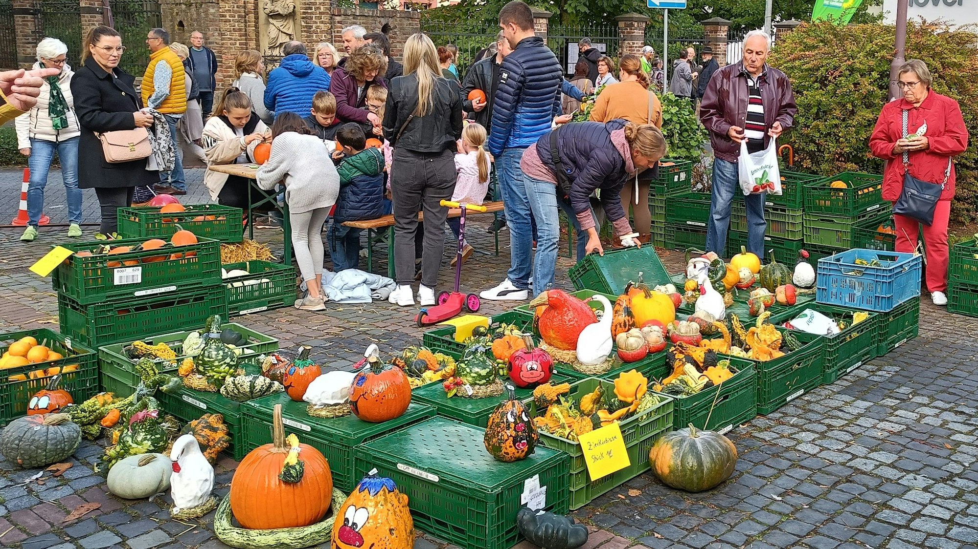 Um einen Obststand mit vielen bunten Kürbissen in Kisten stehen Menschen, die sich die Waren anschauen oder auf Bierbänken sitzen und die Kürbisse ausschnitzen.