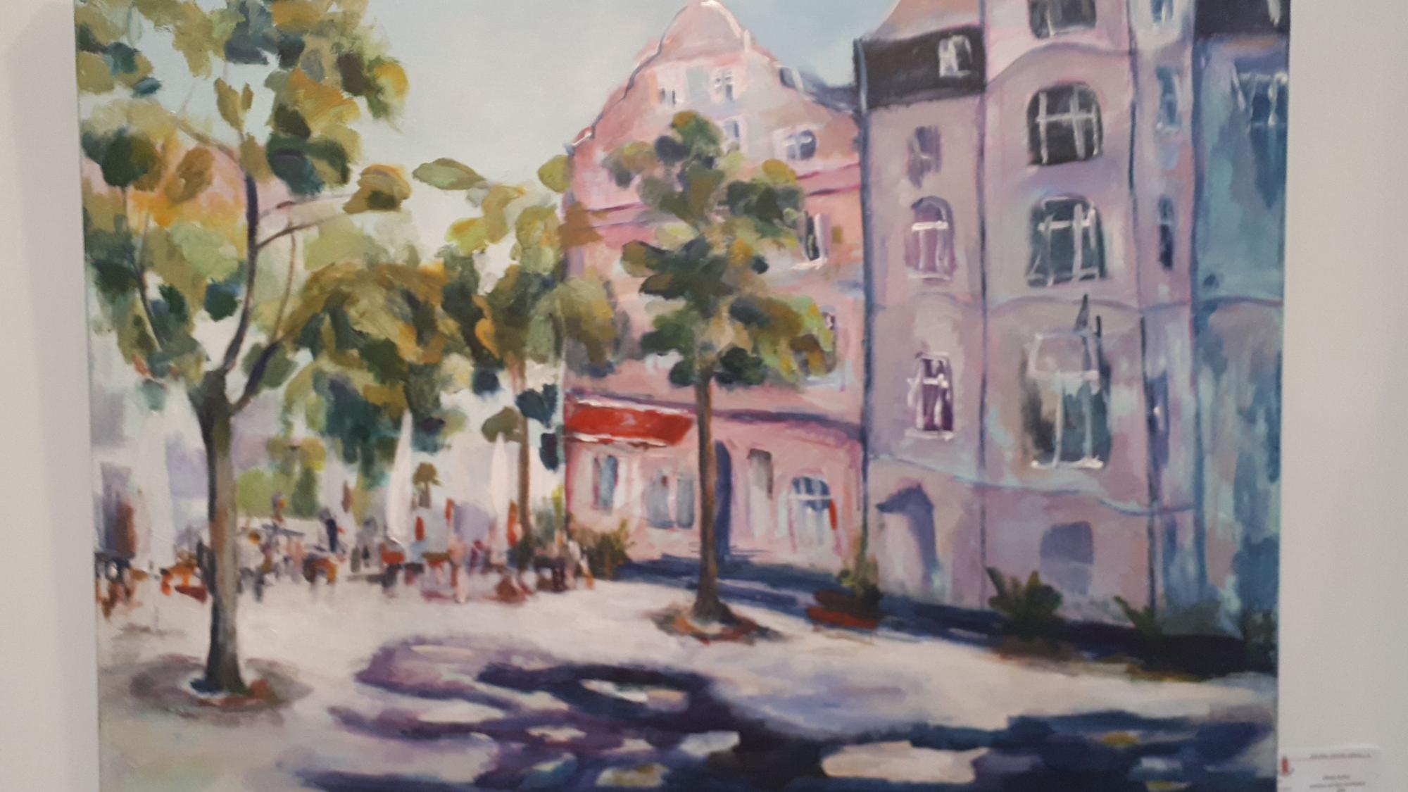 Ein Aquarell vom sommerlichen Schillplatz ist zu sehen. Menschen sitzen draußen an Café-Tischen. Bäume stehen vor den Häusern am beliebten Platz in Nippes.
