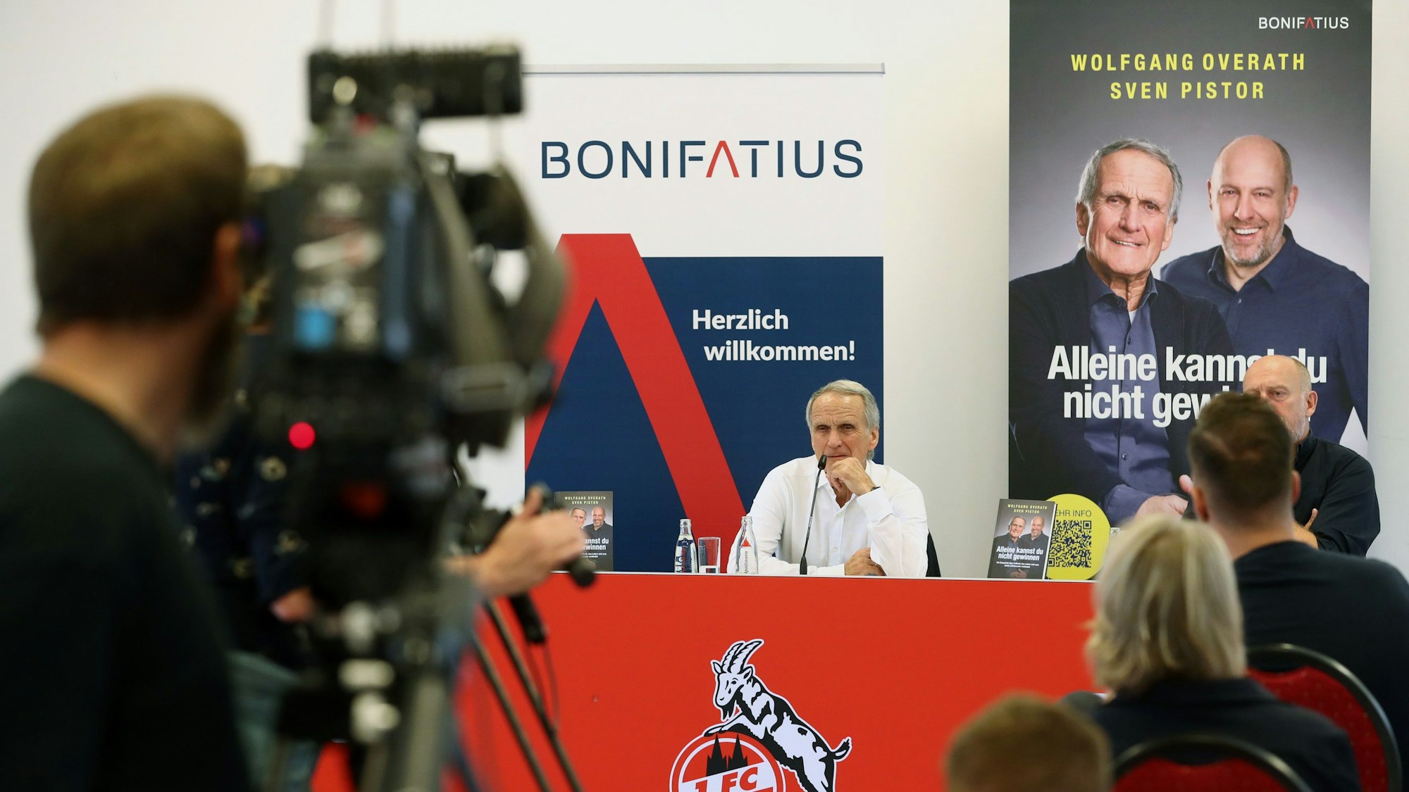 Im Vordergrund steht eine Videokamera und im Hintergrund stellen die Männer ihr Buch vor.