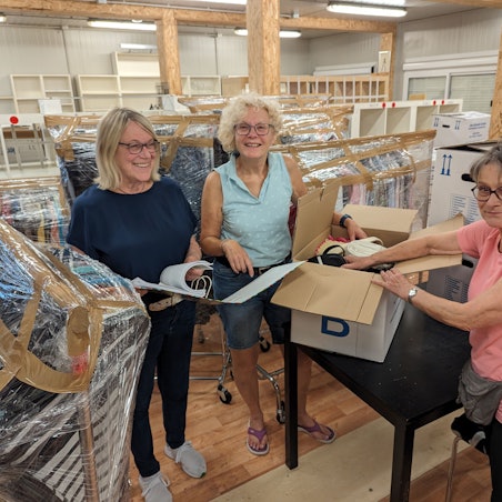 Zwei Frauen packen einen Karton, eine schaut in eine Liste vor leer geräumten Regalen.