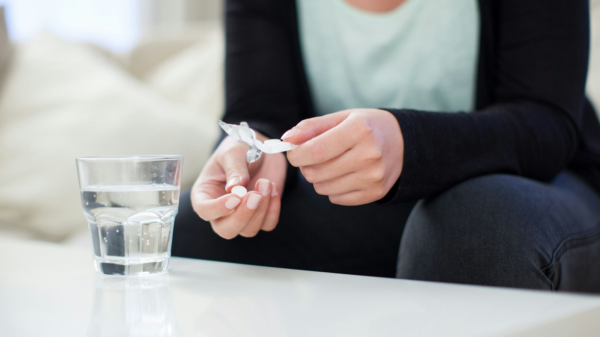 Eine Frau hält eine Tablette und eine Blisterpackung in der Hand. Vor ihr steht ein Glas Wasser.