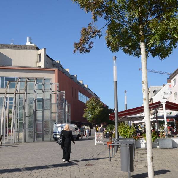 Ein großes Gebäude am Rand einer Fußgängerzone. Am Rand einer gepflasterten Fläche stehen Laternen und einige Bäume.
