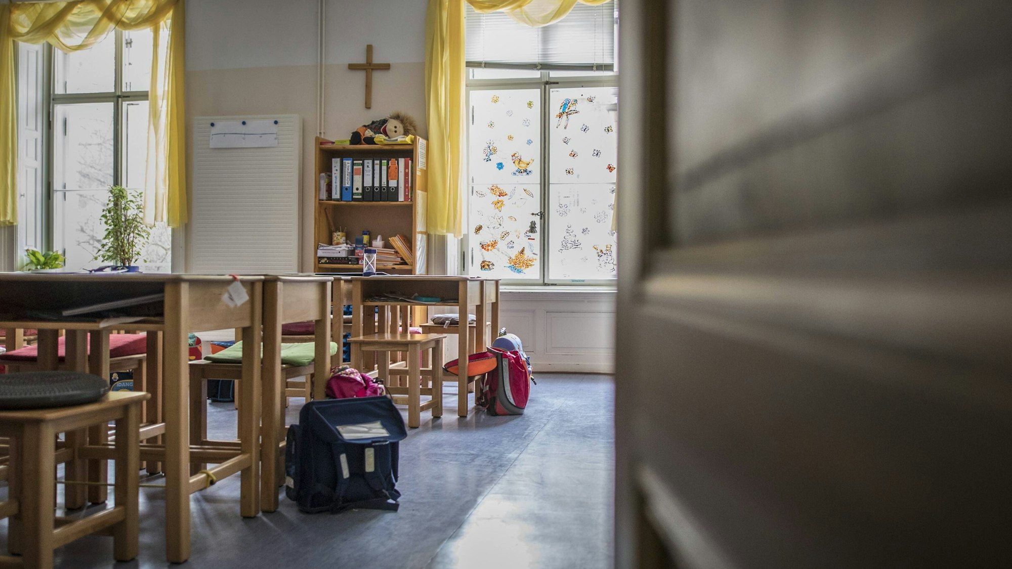 Blick durch eine Tuer in ein leeres Klassenzimmer. An der Wand hängt ein Kreuz.