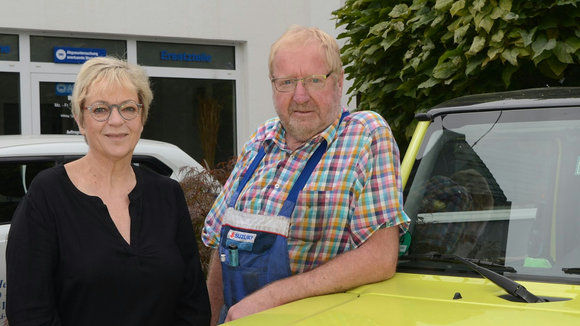 Eine Frau und ein Mann lehnen an der Motorhaube eines gelben Autos. Sie trägt ein schwarzes Oberteil, er ein kariertes Hemd und eine Werkstatt-Latzhose.