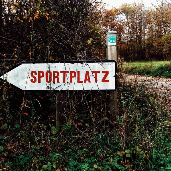 Ein Hinweisschild vor einer Hecke weist den Weg zu einem Sportplatz.