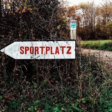 Ein Hinweisschild vor einer Hecke weist den Weg zu einem Sportplatz.