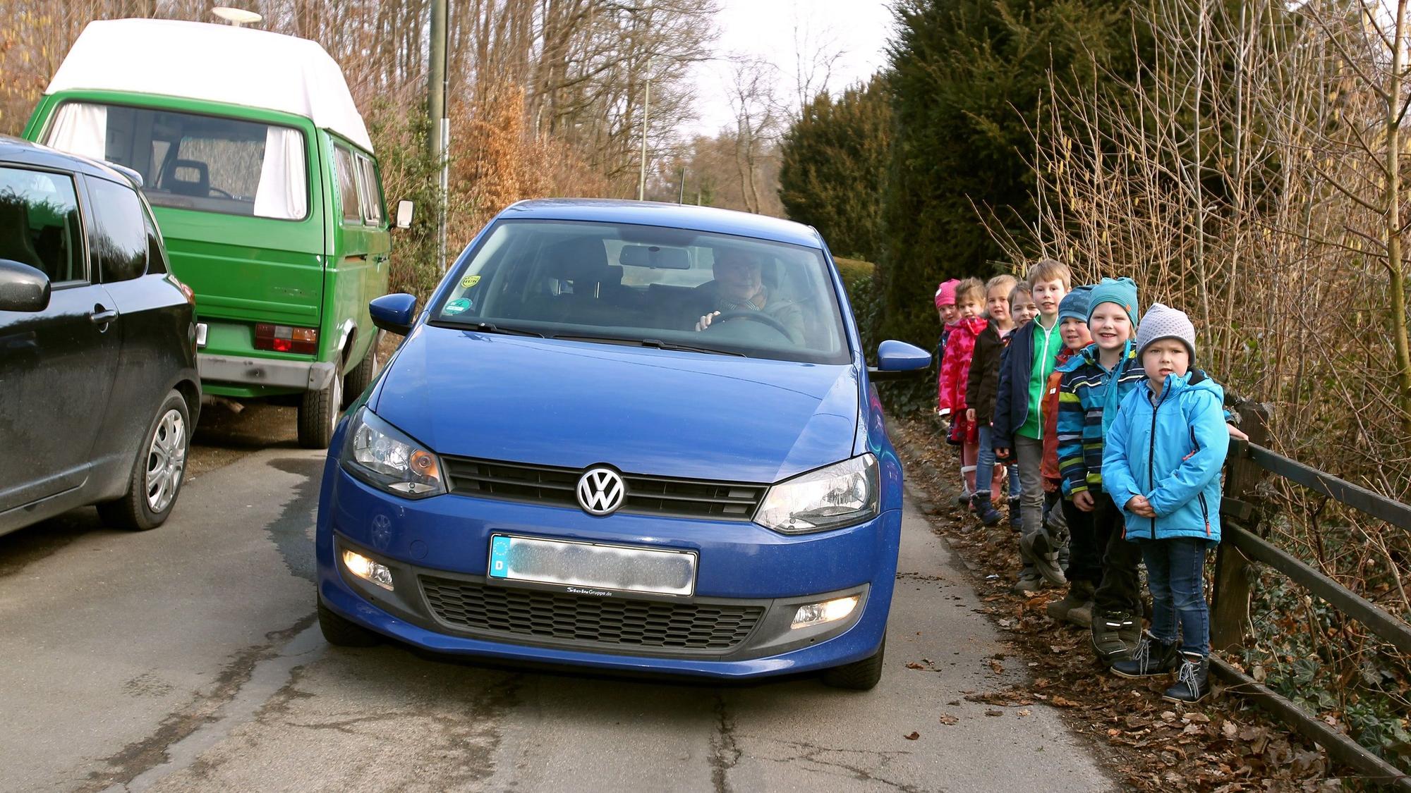 Ein blaues Auto fährt zwischen parkenden Pkw und einer Gruppe von kleinen Kindern hindurch.
