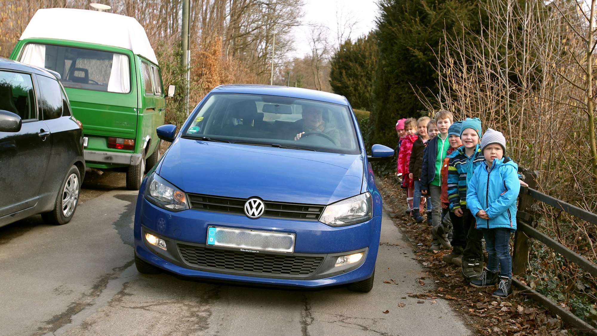 Ein blaues Auto fährt zwischen parkenden Pkw und einer Gruppe von kleinen Kindern hindurch.