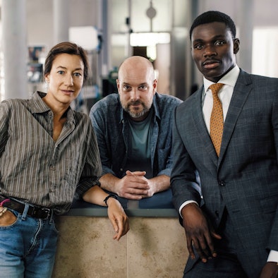 Die drei Schauspieler stehen nebeinander und schauen in die Kamera, im Hintergrund das Büro des Polizeipräsidiums.