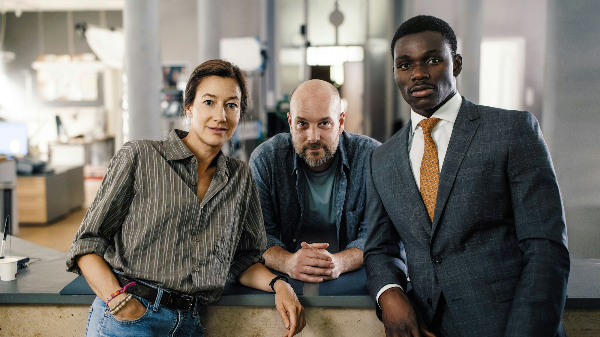 Die drei Schauspieler stehen nebeinander und schauen in die Kamera, im Hintergrund das Büro des Polizeipräsidiums.