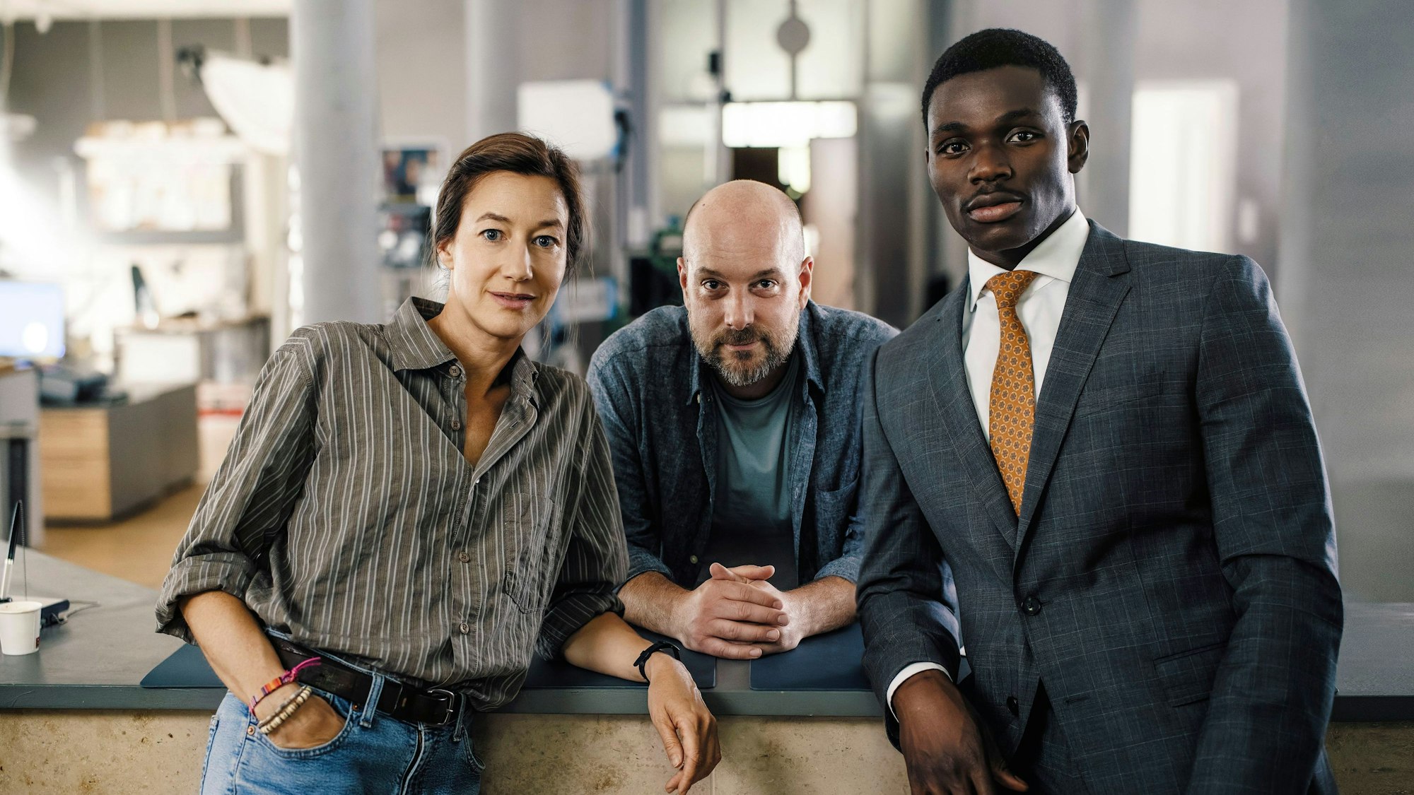 Die drei Schauspieler stehen nebeinander und schauen in die Kamera, im Hintergrund das Büro des Polizeipräsidiums.