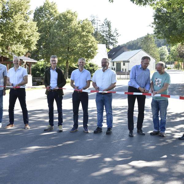 Acht Männer stehen auf einer Straße und halten Scheren sowie rot-weißes Flatterband in den Händen. Links und rechts stehen Bäume. Im Hintergrund sind Häuser zu erkennen.