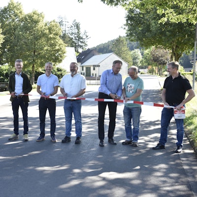 Acht Männer stehen auf einer Straße und halten Scheren sowie rot-weißes Flatterband in den Händen. Links und rechts stehen Bäume. Im Hintergrund sind Häuser zu erkennen.