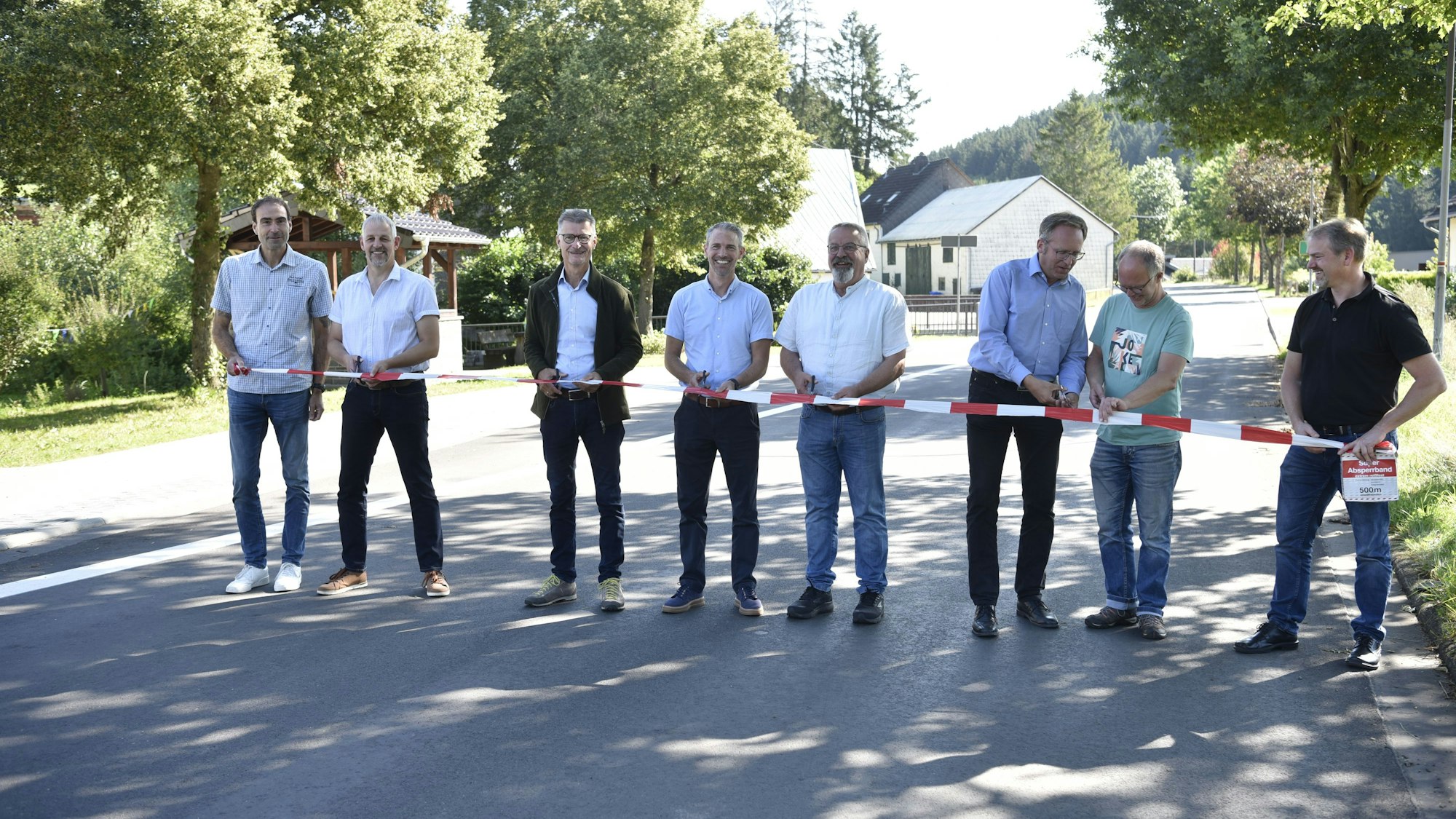 Acht Männer stehen auf einer Straße und halten Scheren sowie rot-weißes Flatterband in den Händen. Links und rechts stehen Bäume. Im Hintergrund sind Häuser zu erkennen.