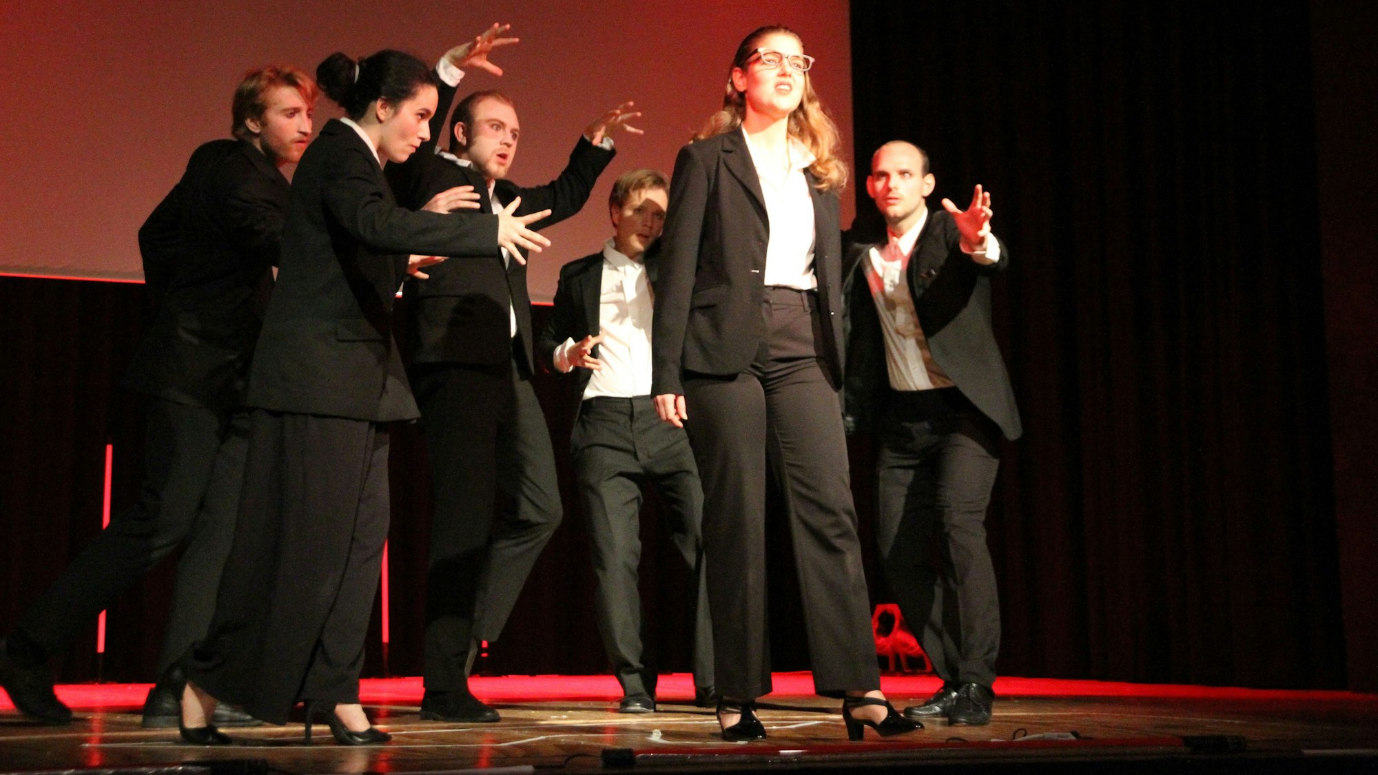 Sieben Schauspieler uns Schauspielerinnen in dunklen Kostümen auf der Bühne in der Aula des Gymnasiums Alleestraße