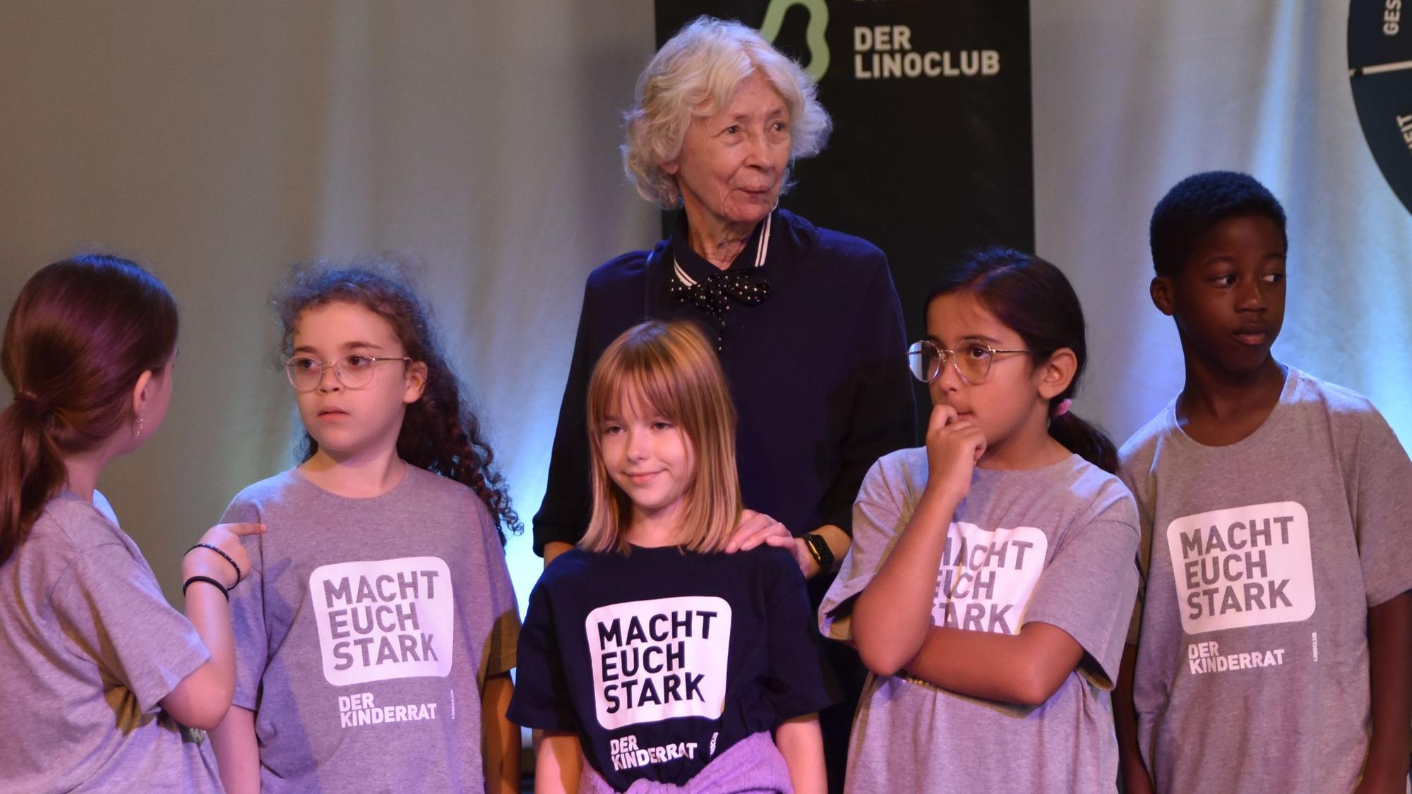 Hedwig Neven DuMont sthet mit fünf jungen Mitgliedern des „Linoclub“-Kinderrats auf der Bühne bei der Generalprobe für das Bühnenprogramm des Weltkindertags. Die Kinder tragen T-Shirts mit der Aufschrift: „Macht euch stark!“
