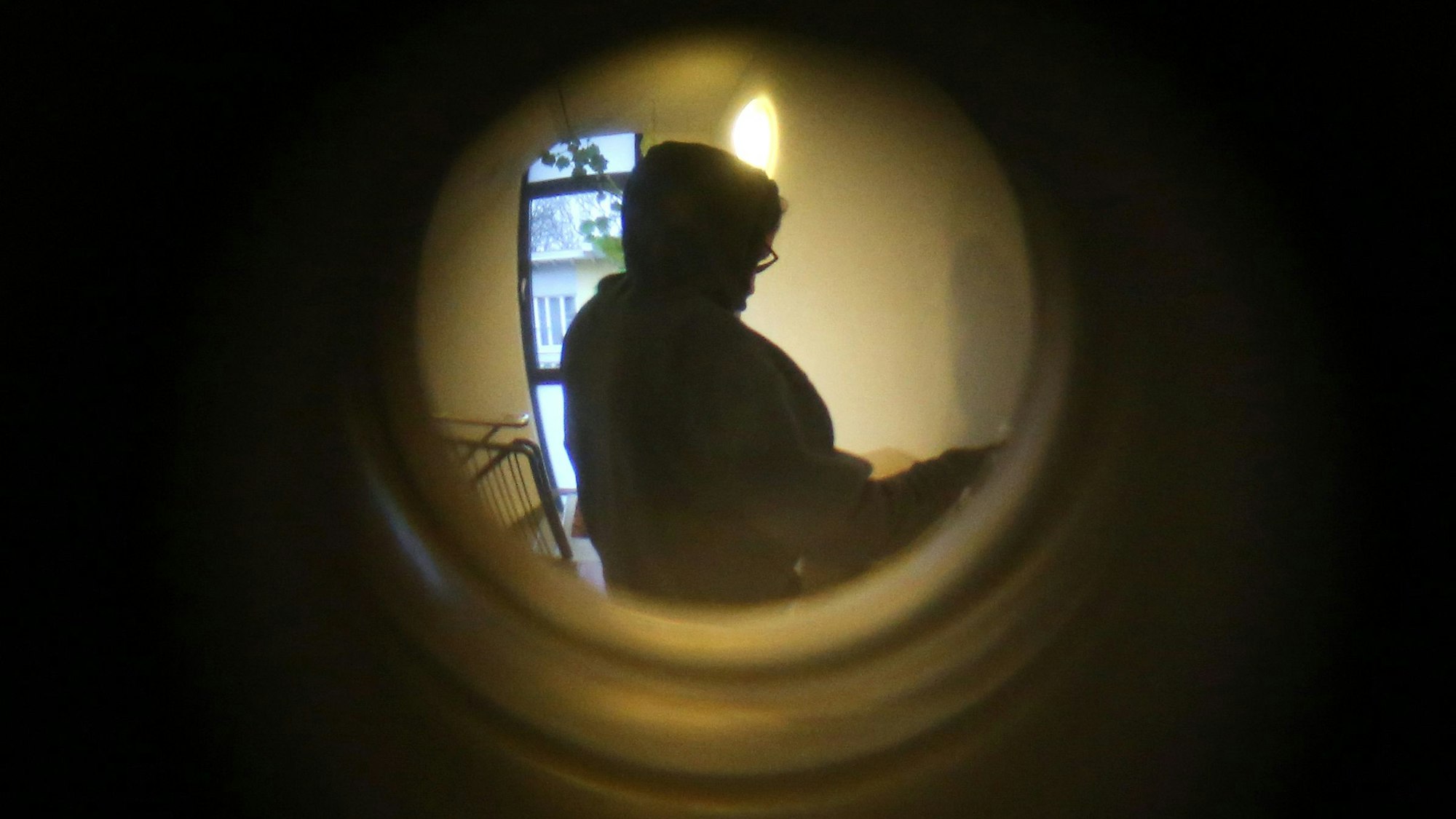 Durch einen Türspion ist eine Person mit einem Kapuzenpulli vor einer Wohnungstür zu sehen.