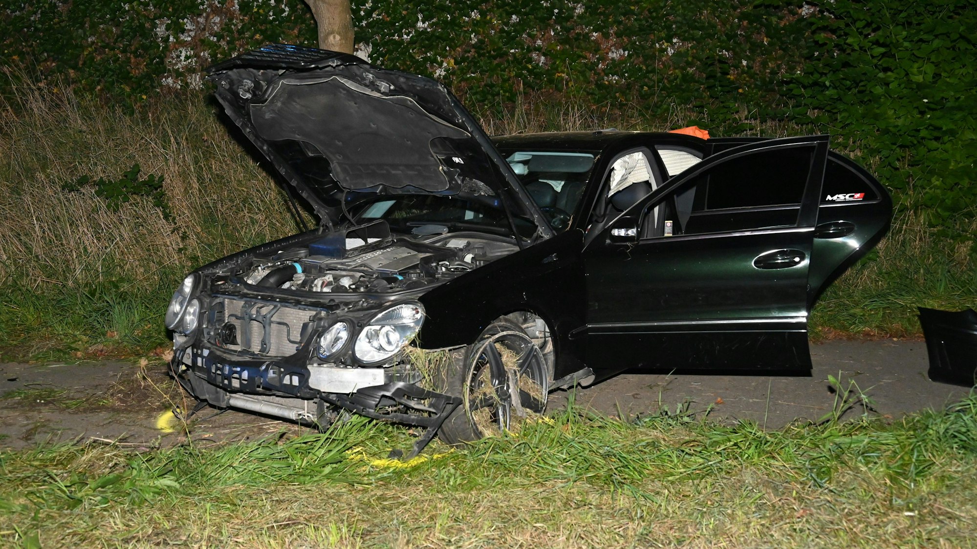 Der beschädigte schwarze Mercedes mit offener Motorhaube und offenen Türen.