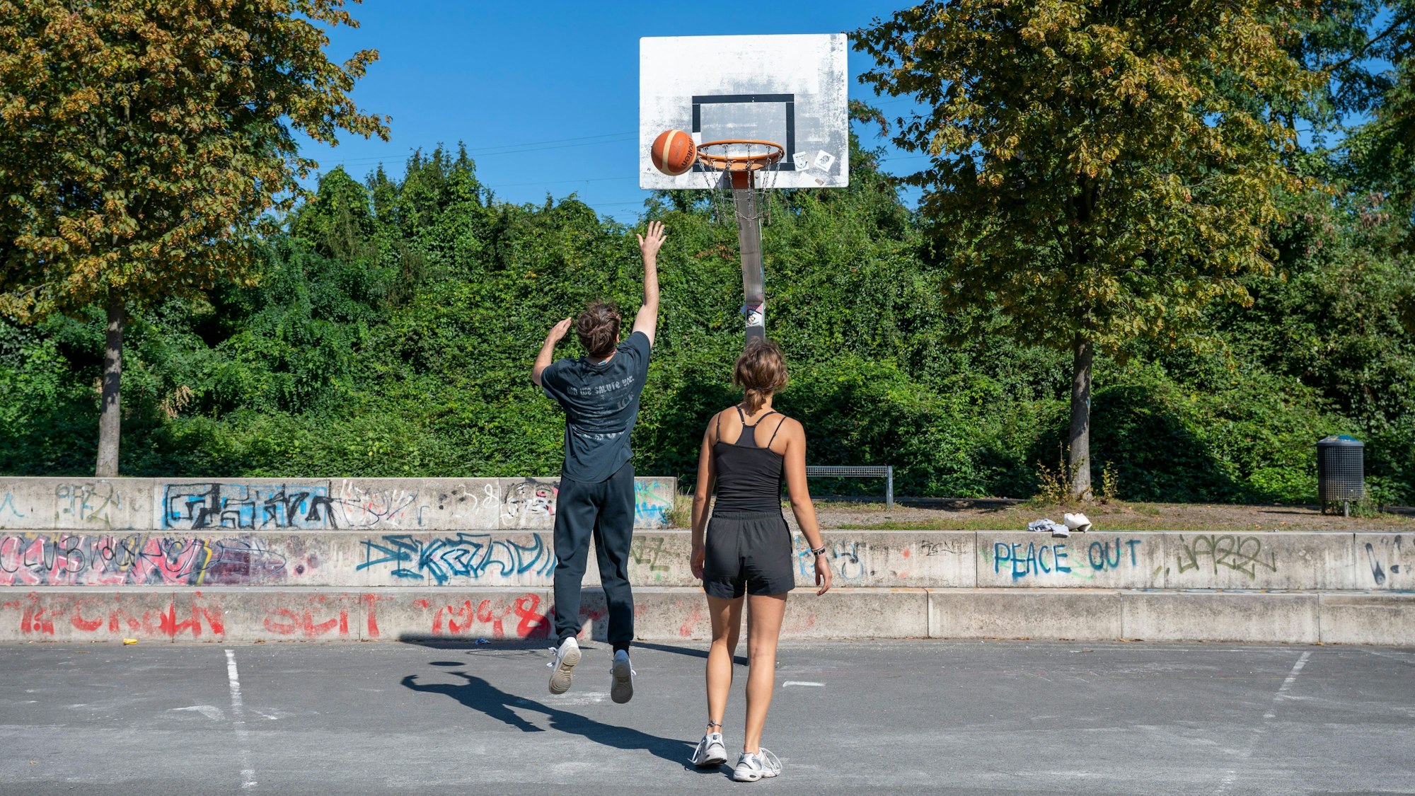 Ein Junge und eine Frau spielen Basketball auf einem Platz mit Bäumen im Hintergrund.