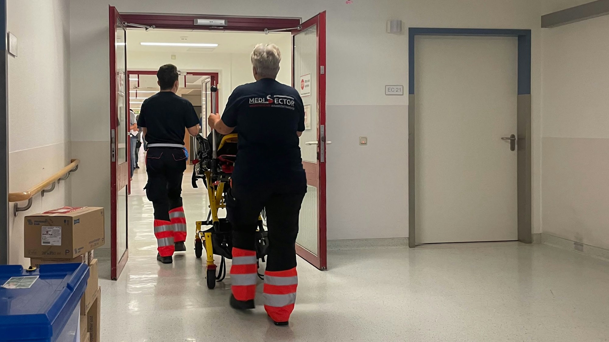 Zwei Rettungssanitäter schieben einen Patienten (verdeckt) auf einer Liege durch die Tür der Notaufnahme.