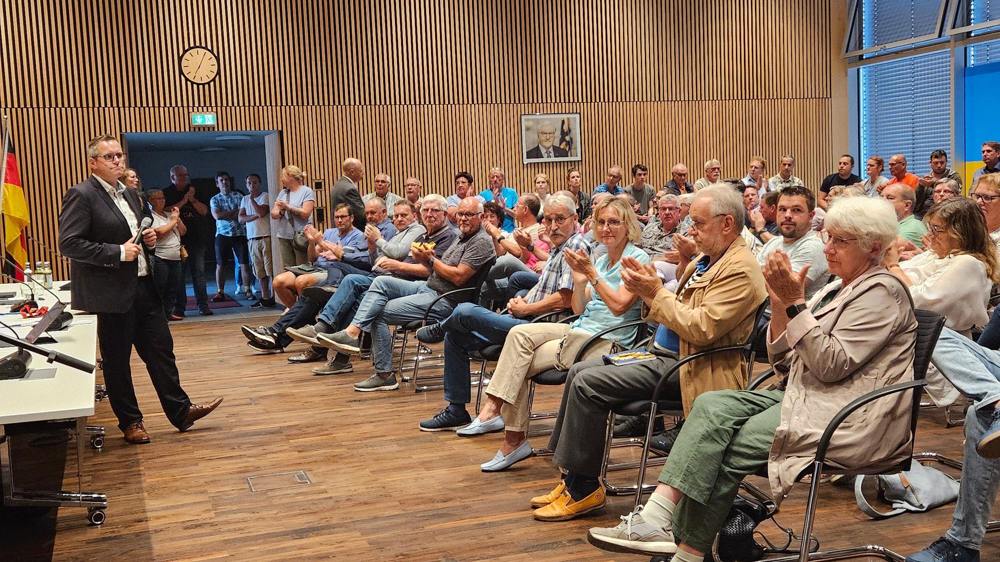 Ein Mann steht in einem großen Saal vor vielen Menschen, die in mehreren Stuhlreihen sitzen und applaudieren.