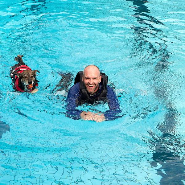 Ein Mann mit langer Schwimmkleidung und Schwimmweste schwimmt neben einem Hund in roter Schwimmweste im Becken eines Freibades.