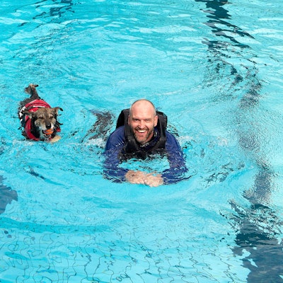 Ein Mann mit langer Schwimmkleidung und Schwimmweste schwimmt neben einem Hund in roter Schwimmweste im Becken eines Freibades.