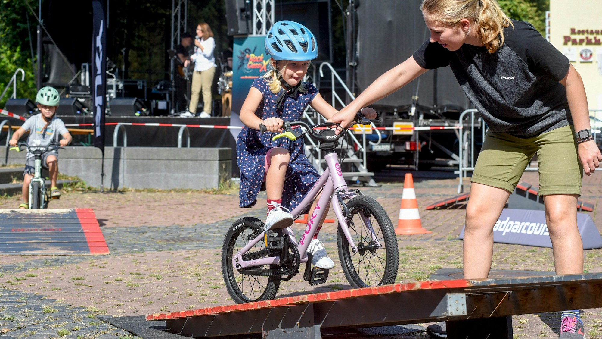 Ein Mädchen fährt mit einem Fahrrad auf eine Rampe, eine junge Frau hilft ihm.