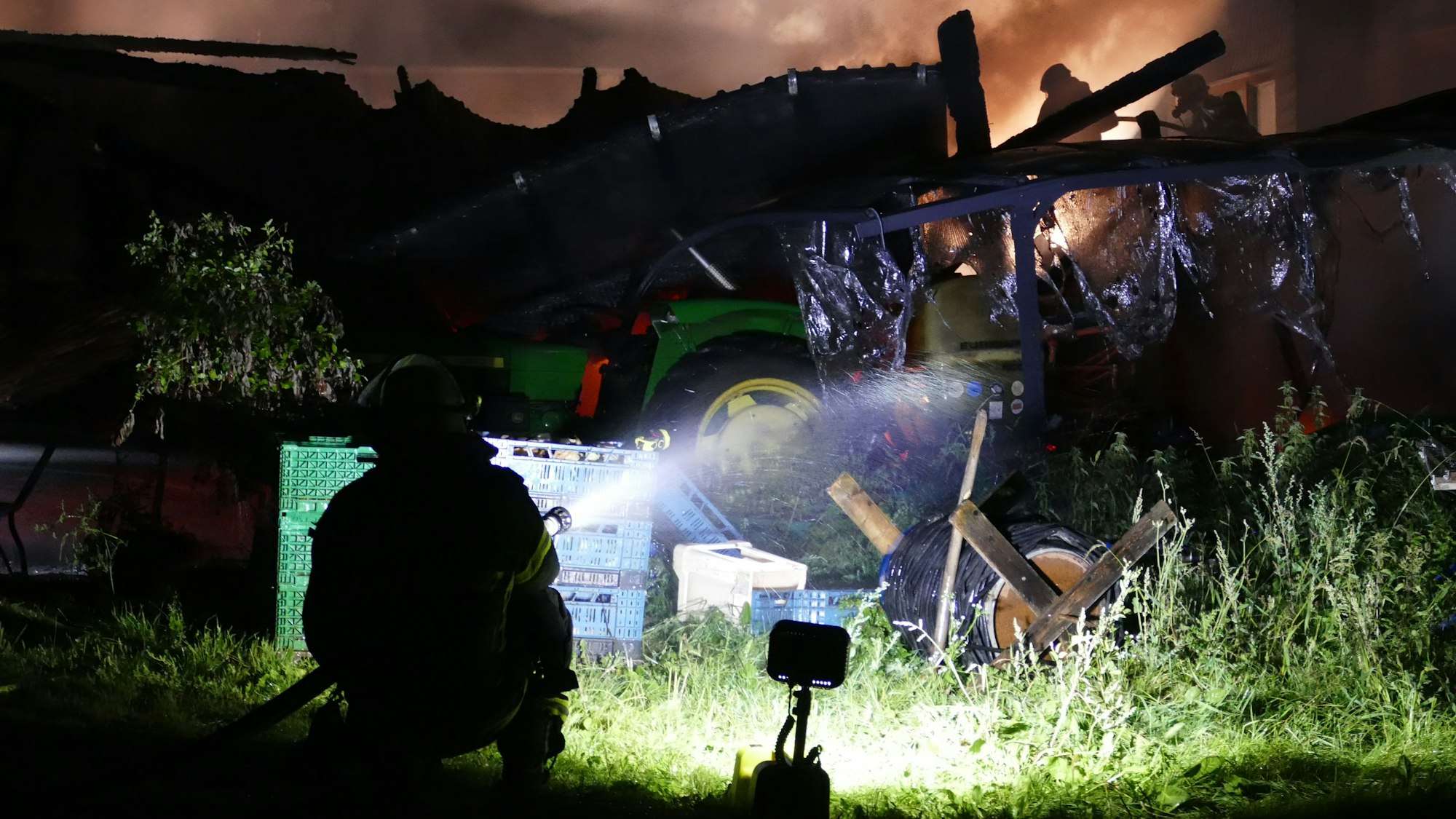 Ein Feuerwehrmann hockt auf dem Boden, im Hintergrund sieht man die eingestürzte Scheune.