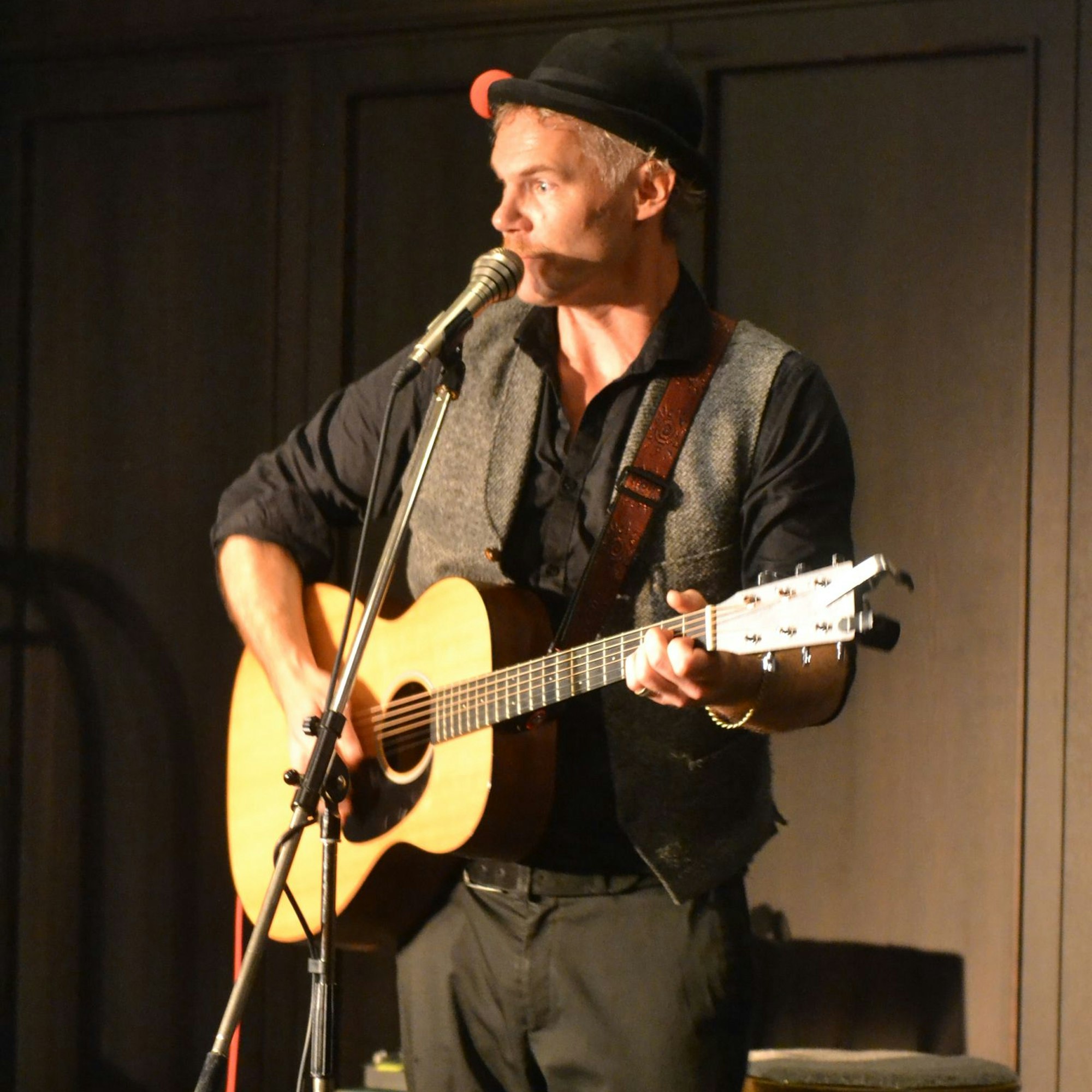 Nisse Barfuss steht mit einer Gitarre an einem Mikrofon, er trägt schwarze Kleidung und einen Hut.