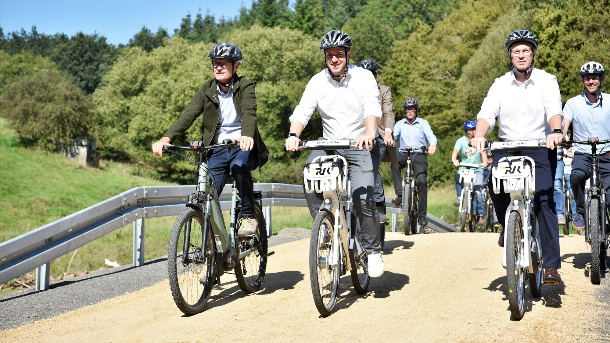 Die Politiker fahren mit dem Rad über einen Weg. Im Hintergrund sind Bäume zu erkennen.