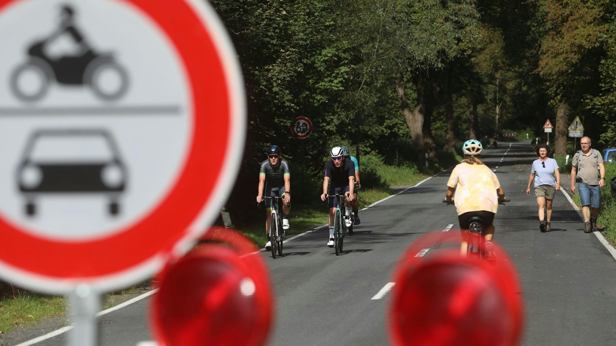 Hinter einem Verbotsschild für Motorräder und Autos sieht man Radfahrer und Wanderer auf einer langen, baumgesäumten Straße.